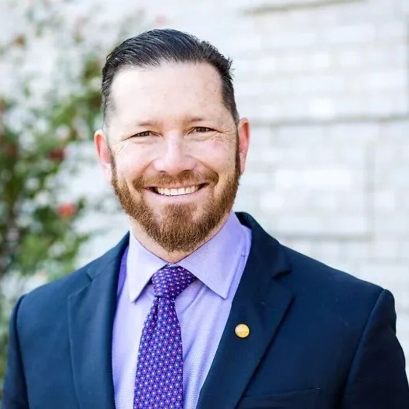 The image shows a man with short brown hair and a neatly trimmed beard. He is wearing a navy blue suit, a light purple shirt, and a purple patterned tie. He has a warm smile and appears to be looking directly at the camera. The background is blurred, but it seems to be an outdoor setting.